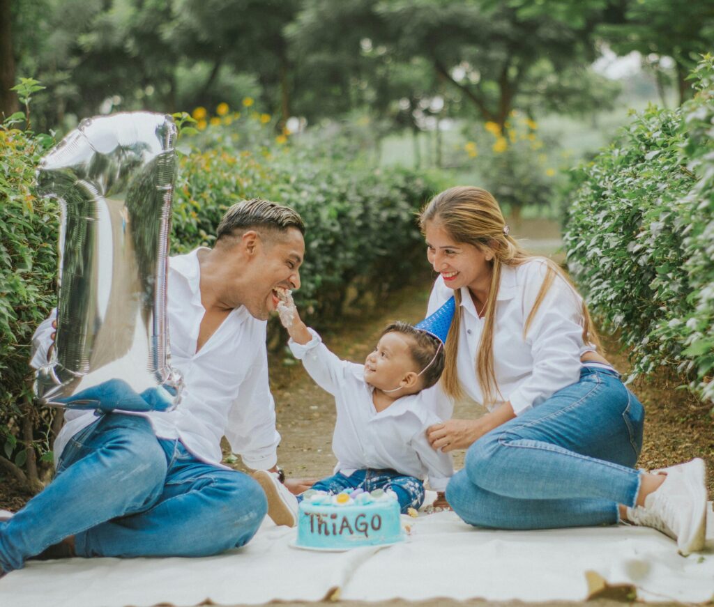 A family celebration in a garden, featuring a child in a birthday hat reaching for a blue cake decorated with colorful sweets.