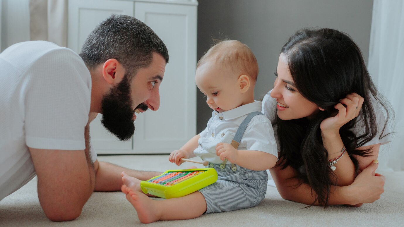 A toddler sits on the floor playing with a colorful toy, while two adults lean in attentively, creating a warm family moment.