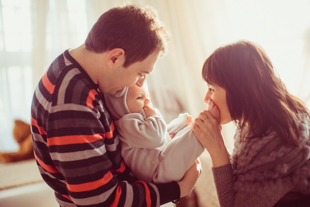 A parent holds a baby dressed in a cozy onesie, while another parent gently interacts with the child in a warmly lit room.