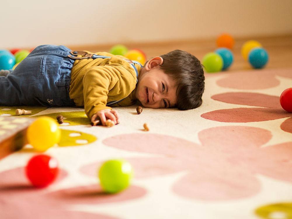 A toddler in a yellow sweater and overalls lays on a colorful rug, surrounded by vibrant plastic balls and wooden toys.