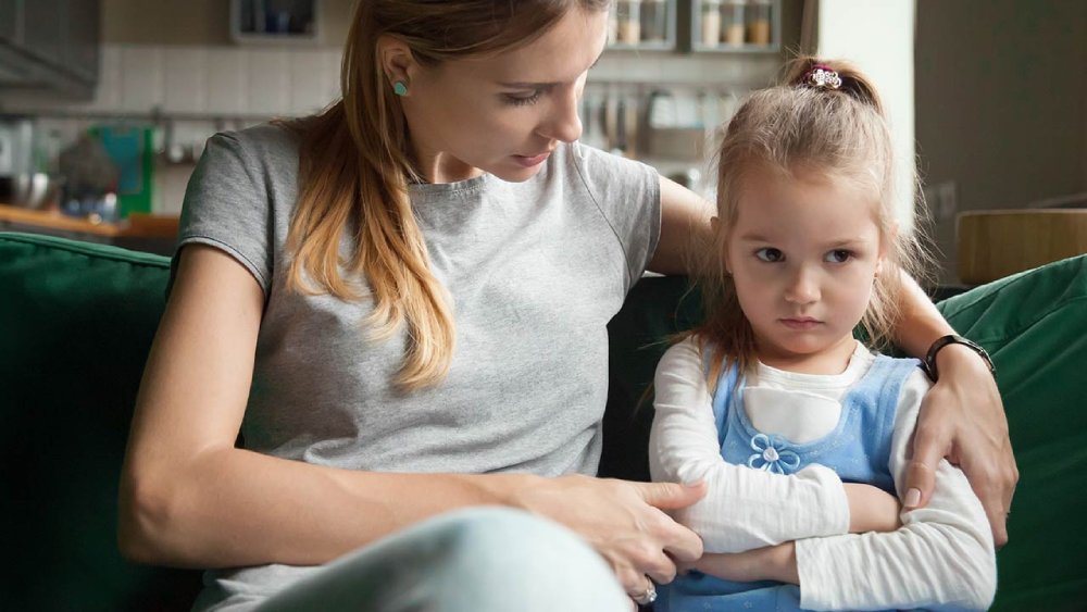 A woman in a gray t-shirt sits on a green couch with a young girl in a blue dress, both relaxed in a cozy home setting.