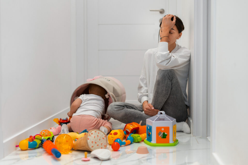 A child plays with scattered colorful toys on a shiny floor while an adult sits against the wall, looking overwhelmed in a hallway.